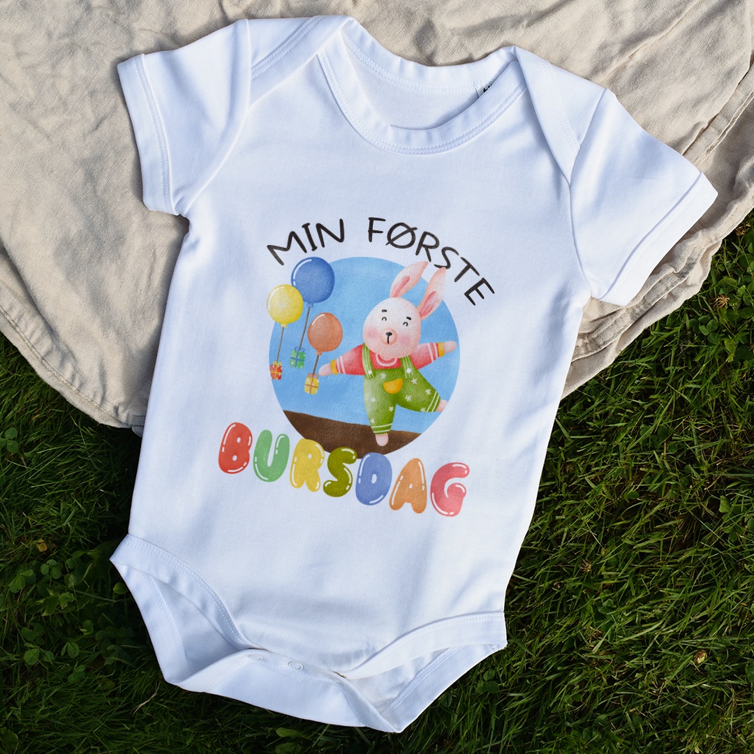 Hvit babybody som ligger flatt delvis på et teppe og delvis på gress, med teksten "Min første bursdag" og figur av en glad kanin og gaver som blir holdt oppe i luften av ballonger. Foto.
