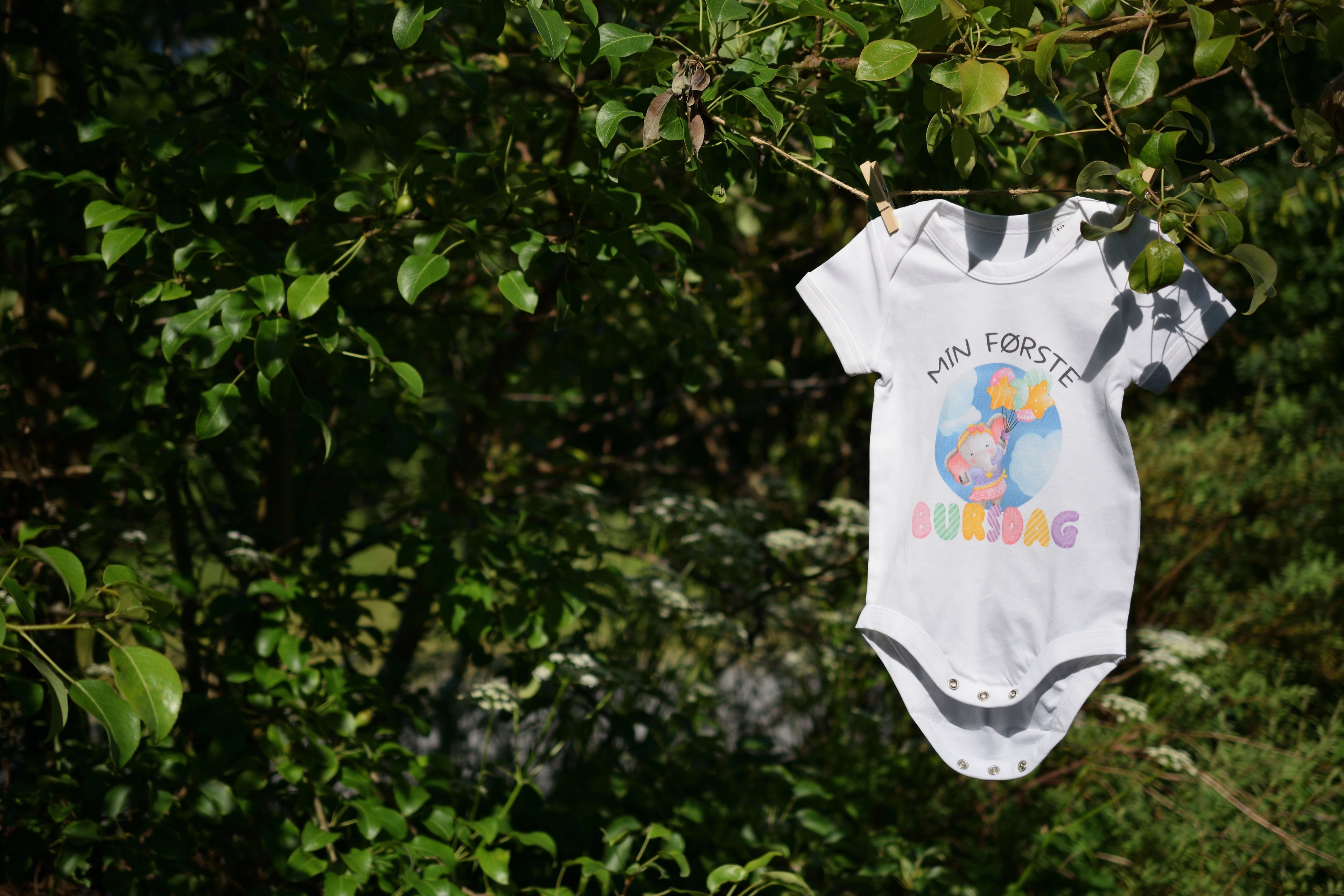 En hvit "Min første bursdag" body med elefant hengt opp på en klessnor ute om sommeren. I bakgrunnen er det mye grønnfarge av blader fra trær. Foto.