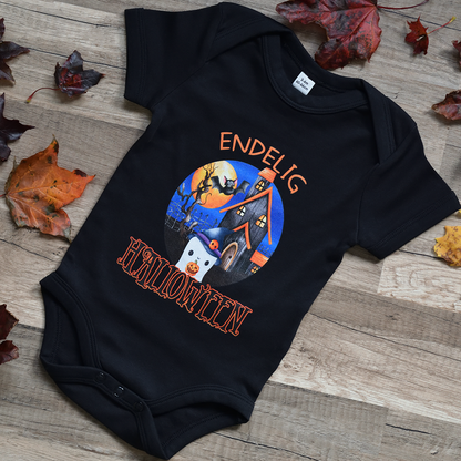 Svart babybody med teksten "Endelig halloween" og figur av et søtt spøkelse med heksehatt foran et hus med en stor fullmåne i bakgrunnen. Bodyen ligger flatt på en trebakgrunn, med høstblader rundt. Foto.