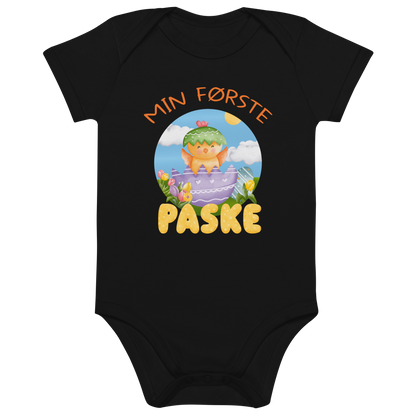 Svart babybody som ligger flatt, med teksten "Min første påske" og figur av en glad kylling som hopper ut av et påskeegg. Foto.