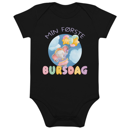 Svart babybody som ligger flatt, med teksten "Min første bursdag" og figur av en søt og glad elefant som holder flere ballonger og flyr mellom skyene. Foto.