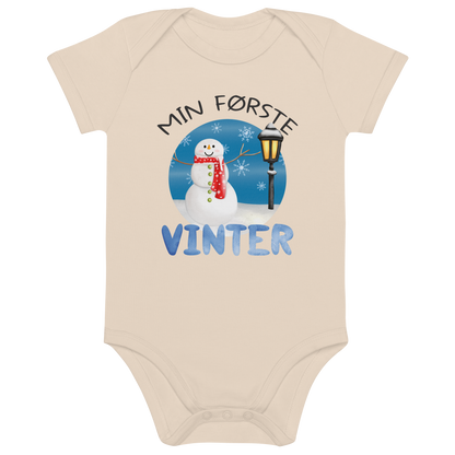 Beige babybody som ligger flatt, med teksten "Min første vinter" og figur av en glad snømann ved en gammeldags lyktestolpe. Foto.
