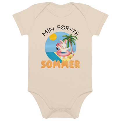 Beige babybody som ligger flatt, med teksten "Min første sommer" og figur av en glad kanin med flamingo badering på stranden. Foto.