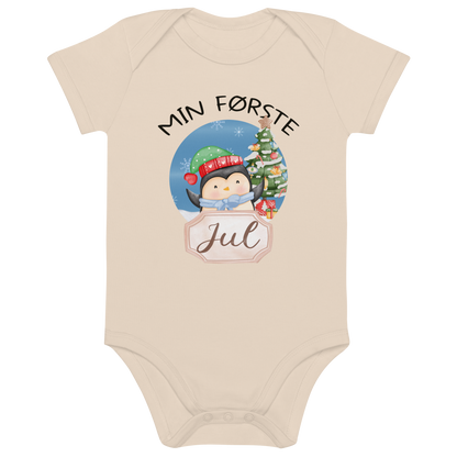Beige babybody som ligger flatt, med teksten "Min første jul" og figur av en glad pingvin med nisselue og skjerf foran et juletre. Foto.
