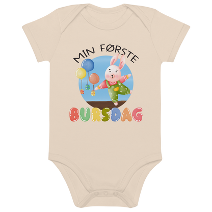 Beige babybody som ligger flatt, med teksten "Min første bursdag" og figur av en glad kanin og gaver som blir holdt oppe i luften av ballonger. Foto.