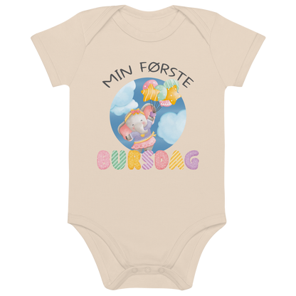 Beige babybody som ligger flatt, med teksten "Min første bursdag" og figur av en søt og glad elefant som holder flere ballonger og flyr mellom skyene. Foto.