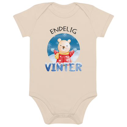 Beige babybody som ligger flatt, med teksten "Endelig vinter" og figur av en lykkelig bamse i vinterklær som nyter snøen. Foto.