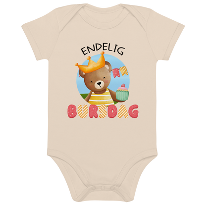Beige babybody som ligger flatt, med teksten "Endelig bursdag" og figur av en glad bamse som har på seg en krone og holder frem en muffin. Foto.