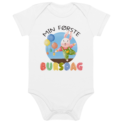 Hvit babybody som ligger flatt, med teksten "Min første bursdag" og figur av en glad kanin og gaver som blir holdt oppe i luften av ballonger. Foto.