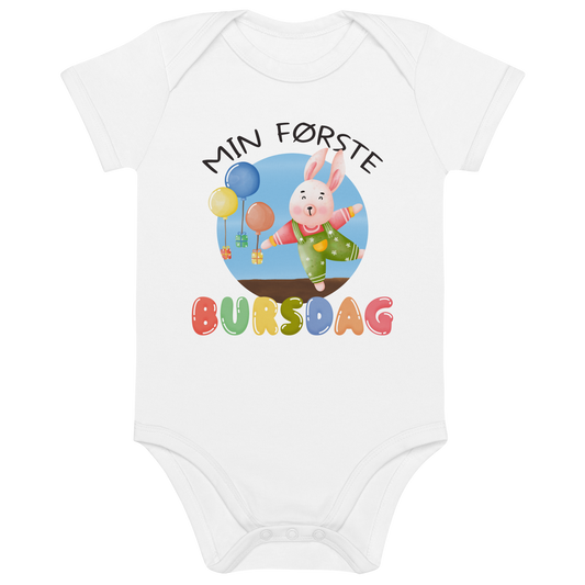 Hvit babybody som ligger flatt, med teksten "Min første bursdag" og figur av en glad kanin og gaver som blir holdt oppe i luften av ballonger. Foto.