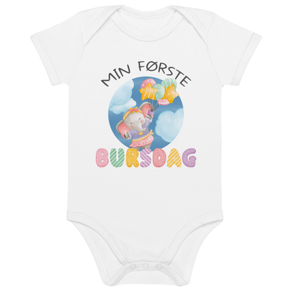 Hvit babybody som ligger flatt, med teksten "Min første bursdag" og figur av en søt og glad elefant som holder flere ballonger og flyr mellom skyene. Foto.