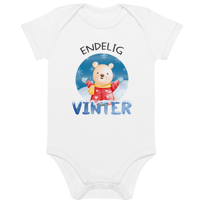 Hvit babybody som ligger flatt, med teksten "Endelig vinter" og figur av en lykkelig bamse i vinterklær som nyter snøen. Foto.
