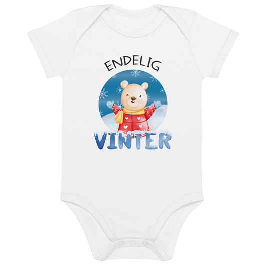 Hvit babybody som ligger flatt, med teksten "Endelig vinter" og figur av en lykkelig bamse i vinterklær som nyter snøen. Foto.