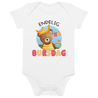 Hvit babybody som ligger flatt, med teksten "Endelig bursdag" og figur av en glad bamse som har på seg en krone og holder frem en muffin. Foto.
