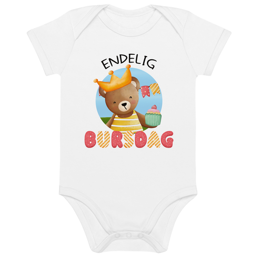 Hvit babybody som ligger flatt, med teksten "Endelig bursdag" og figur av en glad bamse som har på seg en krone og holder frem en muffin. Foto.