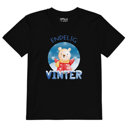 Svart t-skjorte som ligger flatt, med teksten "Endelig vinter" og figur av en lykkelig bamse i vinterklær som nyter snøen. Foto.