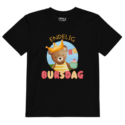 Svart t-skjorte til barn som ligger flatt, med teksten "Endelig bursdag" og figur av en glad bamse som har på seg en krone og holder frem en muffin. Foto.