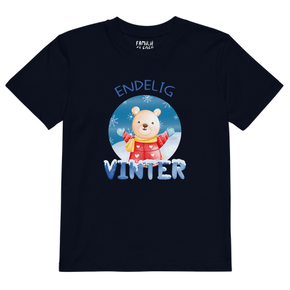 Mørkeblå t-skjorte som ligger flatt, med teksten "Endelig vinter" og figur av en lykkelig bamse i vinterklær som nyter snøen. Foto.