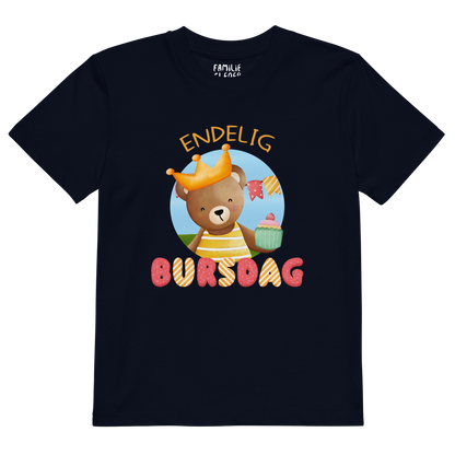 Mørkeblå t-skjorte til barn som ligger flatt, med teksten "Endelig bursdag" og figur av en glad bamse som har på seg en krone og holder frem en muffin. Foto.