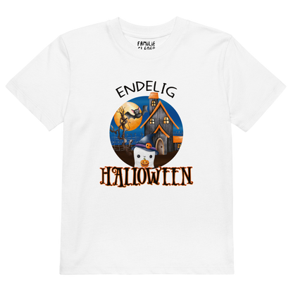Hvit t-skjorte som ligger flatt, med teksten "Endelig halloween" og figur av et søtt spøkelse med heksehatt foran et hus med en stor fullmåne i bakgrunnen. Foto.