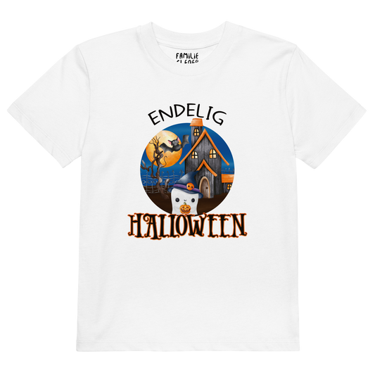 Hvit t-skjorte som ligger flatt, med teksten "Endelig halloween" og figur av et søtt spøkelse med heksehatt foran et hus med en stor fullmåne i bakgrunnen. Foto.