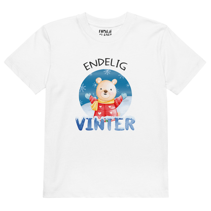 Hvit t-skjorte som ligger flatt, med teksten "Endelig vinter" og figur av en lykkelig bamse i vinterklær som nyter snøen. Foto.