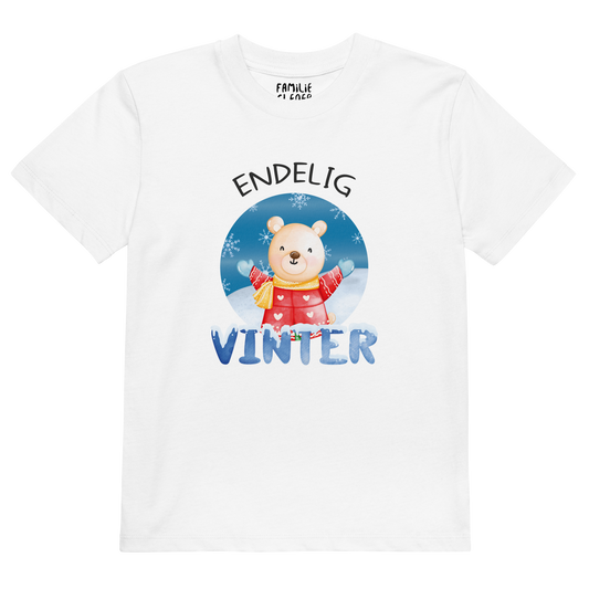 Hvit t-skjorte som ligger flatt, med teksten "Endelig vinter" og figur av en lykkelig bamse i vinterklær som nyter snøen. Foto.