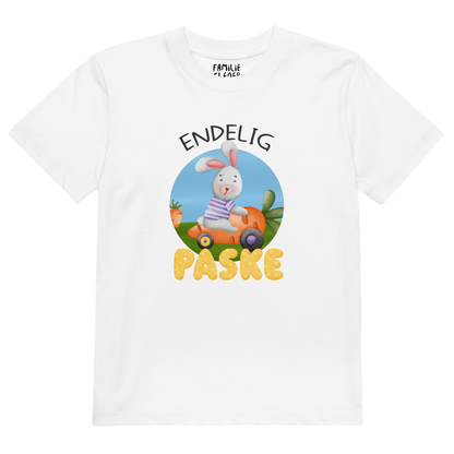 Hvit t-skjorte som ligger flatt, med teksten "Endelig påske" og figur av en glad kanin som kjører en gulrotbil. Foto.