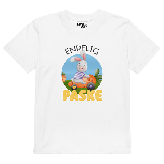 Hvit t-skjorte som ligger flatt, med teksten "Endelig påske" og figur av en glad kanin som kjører en gulrotbil. Foto.