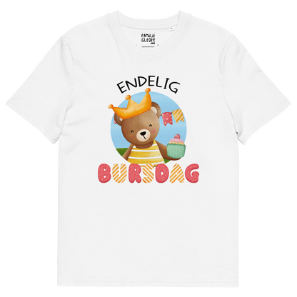Hvit t-skjorte som ligger flatt, med teksten "Endelig bursdag" og figur av en glad bamse som har på seg en krone og holder frem en muffin. Foto.