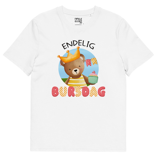Hvit t-skjorte som ligger flatt, med teksten "Endelig bursdag" og figur av en glad bamse som har på seg en krone og holder frem en muffin. Foto.